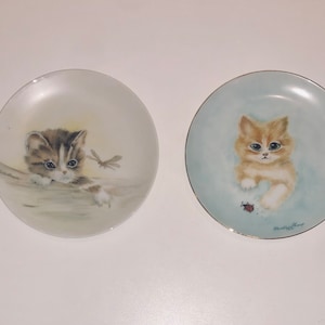 May include: Two decorative ceramic plates featuring images of kittens. The plate on the left depicts a tabby kitten with a dragonfly, while the plate on the right shows a ginger kitten with a ladybug.