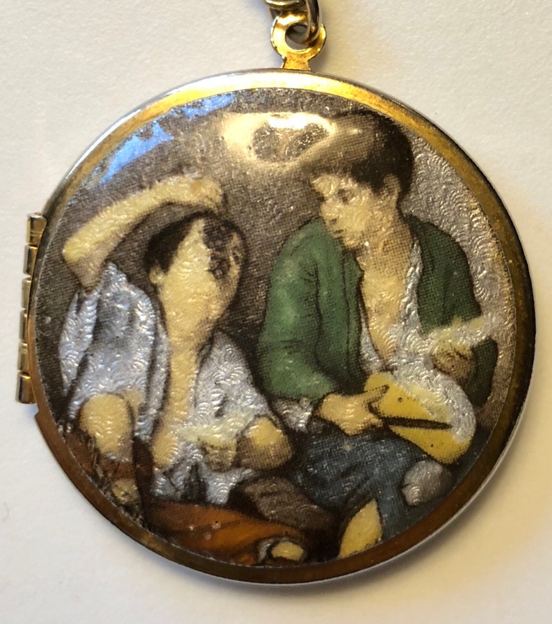 May include: A round, gold-toned locket with a painted scene of two figures. One figure wears a green jacket, the other a white shirt. The locket is attached to a gold chain.