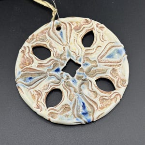 May include: White ceramic ornament with a detailed, embossed design in shades of brown and blue. The ornament has a circular shape with a hole at the top for hanging.