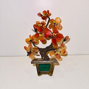 May include: A small, decorative bonsai tree made of red and orange agate stones. The tree is in a green and gold metal pot with a decorative border.