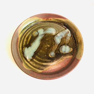 May include: A round ceramic plate with a brown, red and white glaze. The plate has a slightly raised rim and a swirling pattern in the glaze.