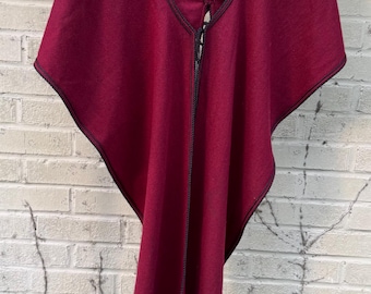 May include: A burgundy poncho with a black trim. The poncho is hanging on a wooden hanger against a white brick wall.