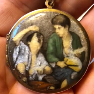 May include: A gold-toned locket pendant with a detailed miniature painting. The painting depicts two figures, one in a green jacket, the other in a white shirt. The locket hangs from a delicate gold chain, showcasing a vintage aesthetic.