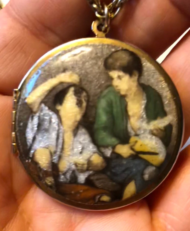 May include: A gold-toned locket pendant with a detailed miniature painting. The painting depicts two figures, one in a green jacket, the other in a white shirt. The locket hangs from a delicate gold chain, showcasing a vintage aesthetic.