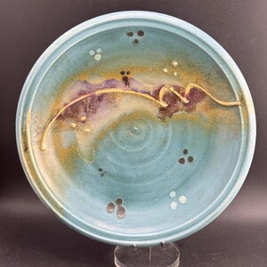 May include: A round ceramic plate featuring a teal and blue glaze with a prominent curved, raised beige line across the center. The surface displays mottled purple and brown accents, along with small clusters of dark and light circular dots.