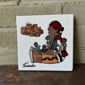 May include: A white ceramic tile with a colorful illustration of a Native American woman holding children. The woman is wearing a red and black dress with turquoise jewelry. The tile also features a small illustration of a pueblo-style building. The tile is signed "Teissedre".