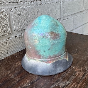 May include: A ceramic pot with a rounded top and a wide base. The pot is glazed in a teal green color with iridescent accents. The base of the pot is a dark gray color.