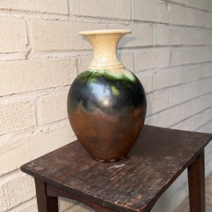 May include: A ceramic vase with a brown, black, and green glaze. The vase has a white rim and a crackled glaze. It is sitting on a wooden table.