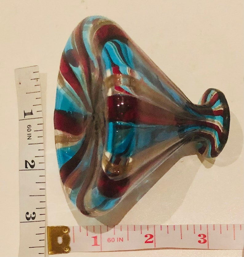 May include: A decorative glass vase with a flared opening and a bulbous body. The vase features a swirl design in shades of blue, red, and gold. The vase is approximately 7.6 cm in height.