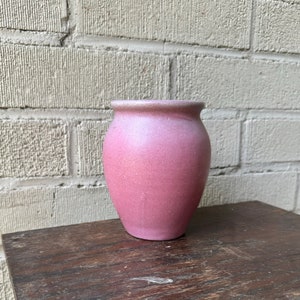 May include: A small, pink ceramic vase with a textured surface. The vase is round and has a slightly flared opening.