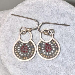 Puede incluir: Un par de pendientes de plata con un pequeño dije redondo de plata. Los dijes están decorados con pequeñas cuentas de colores en tonos rojo, blanco y gris.