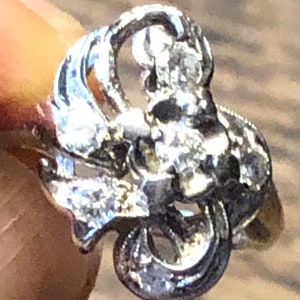 May include: A silver ring with a delicate, intricate design featuring a cluster of small diamonds. The ring has a floral motif and is set in a vintage style.