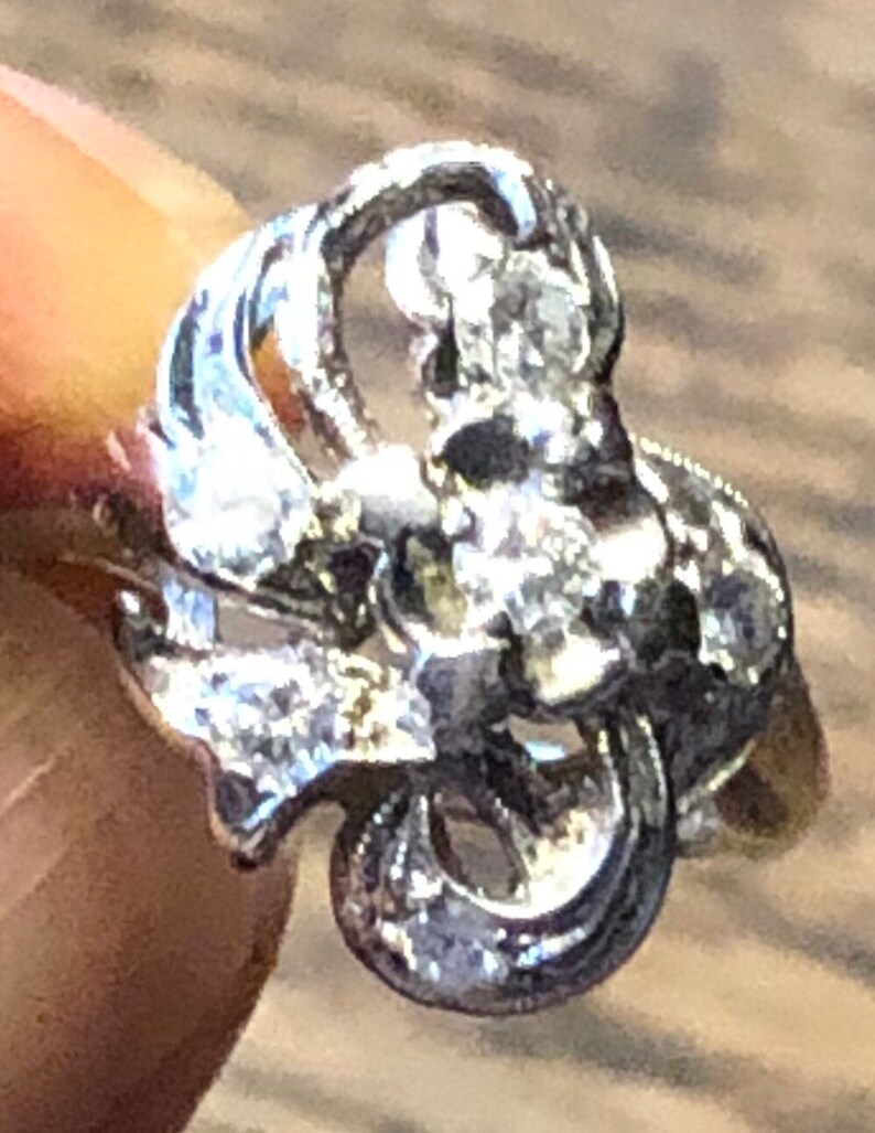 May include: A silver ring with a delicate, intricate design featuring a cluster of small diamonds. The ring has a floral motif and is set in a vintage style.