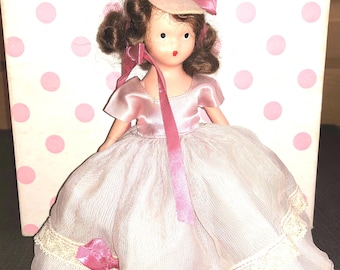 Puede incluir: Una muñeca vintage que lleva un vestido rosa con un borde de encaje blanco y un sombrero rosa con un lazo. La muñeca tiene el pelo castaño y rasgos pintados.