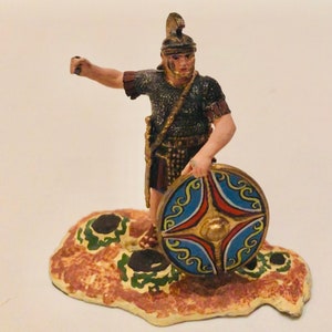 May include: A painted figurine of a Roman soldier wearing a helmet and chainmail, holding a shield with blue, red and gold designs. The soldier is standing on a brown and green base.