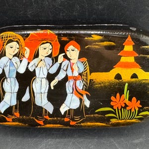 May include: An oval black lacquer box featuring a hand-painted scene of three figures in light blue garments and head coverings walking near a golden pagoda structure and orange flowers. The artwork uses gold, orange, and blue paint accents.