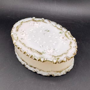 May include: An oval-shaped, white trinket box with a floral design on the lid. The box has a cream-colored base and is trimmed with gold-colored accents. The lid features embossed roses and a scalloped edge.