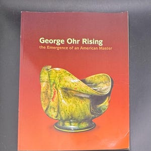 May include: A softcover book titled George Ohr Rising: the Emergence of an American Master. The cover features a deep red to orange gradient background and a photograph of a green and yellow glazed ceramic vessel with a folded, organic shape.