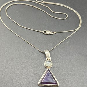 May include: Silver-tone necklace with a triangular pendant. The pendant features a purple gemstone set within a silver frame, topped with a smaller, round, light-colored stone. The chain is a delicate, snake-style design.
