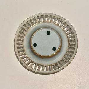 May include: White ceramic saucer with a gold rim and a cut-out design. The saucer has three black rubber feet on the bottom.