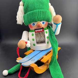 Op de afbeelding: Een houten notenkraker in de vorm van een skiër, met een groene gebreide muts en sjaal. De notenkraker heeft een wit shirt met groene stippen, blauwe ski's en een houten slee. De skiër houdt een houten skistok vast.