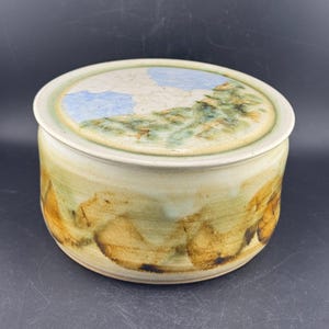 May include: A round ceramic lidded container with a smooth, cylindrical body. The exterior features a blended glaze of earthy brown, muted green, and cream tones. The lid displays a painted landscape scene with blue sky and green foliage.