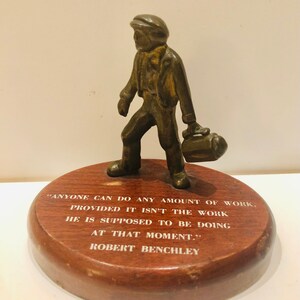May include: A bronze figurine of a man carrying a bag, standing on a wooden base with the quote "ANYONE CAN DO ANY AMOUNT OF WORK, PROVIDED IT ISN'T THE WORK HE IS SUPPOSED TO BE DOING AT THAT MOMENT." by Robert Benchley.