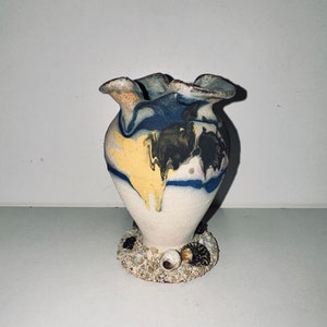 Pode incluir: Um pequeno vaso de cerâmica branca com um design marmorizado azul, amarelo e castanho. O vaso tem um topo franzido e uma base decorada com pequenas conchas.