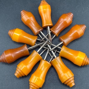 Op de afbeelding: Een verzameling van acht maïskolfhouders gerangschikt in een cirkelvormig patroon. Elke houder heeft een glad, barnsteenkleurig handvat met een getrapt ontwerp aan de basis en twee scherpe, zilverkleurige metalen tanden in het midden.