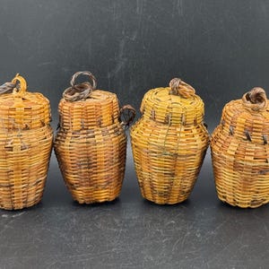 May include: Four small, handwoven baskets with lids. The baskets are made of light brown and dark brown woven material. Each basket has a rounded body and a lid with a small handle. The baskets are arranged in a row against a dark background.