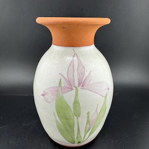 Può includere: Un vaso in ceramica con corpo bianco e bordo color terracotta. Il vaso presenta un disegno dipinto a mano di un fiore di iris rosa con foglie verdi. Il vaso ha una forma arrotondata e un'ampia apertura.