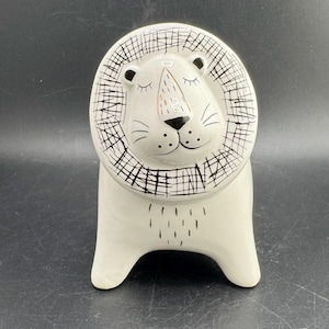 Puede incluir: Una figura de león de cerámica color crema con una melena estilizada de líneas negras entrecruzadas. El león tiene una cara lisa con los ojos cerrados, una nariz negra y pequeños puntos negros que representan los bigotes.