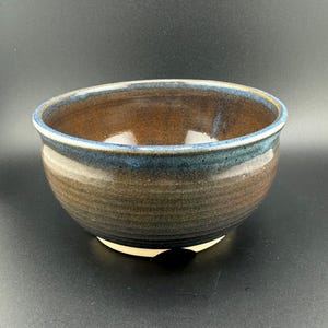 Può includere: Una ciotola in ceramica marrone con una glassa blu. La ciotola ha un fondo leggermente arrotondato e un bordo largo.