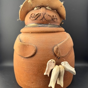 Może przedstawiać: Ceramiczny słoik z terakoty w kształcie kota w kapeluszu z szerokim rondem. Kot ma teksturowaną brązową powierzchnię, obrożę i ramiona złożone na ciele. Z jednego ramienia zwisa drut, na którym zawieszone są cztery małe, kremowobiałe gliniane zawieszki w kształcie ryb.