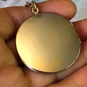 May include: A gold-colored, round locket pendant with a hinged opening. The locket is suspended from a delicate gold chain. The pendant has a smooth, reflective surface, suggesting it may be designed to hold a photograph or small keepsake.