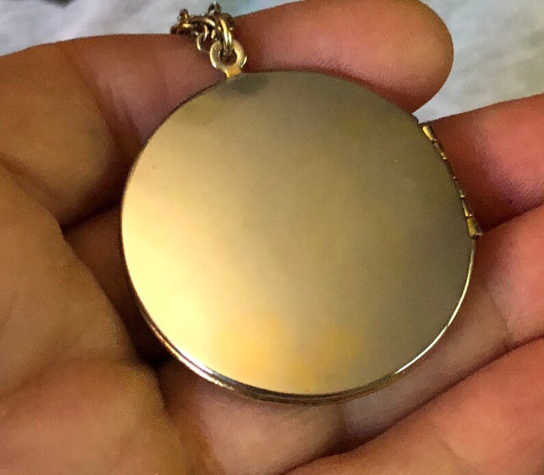 May include: A gold-colored, round locket pendant with a hinged opening. The locket is suspended from a delicate gold chain. The pendant has a smooth, reflective surface, suggesting it may be designed to hold a photograph or small keepsake.