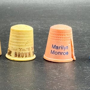 Op de afbeelding: Een set van vier plastic naaivingerhoeden in geel, lichtgeel, perzik en blauw. De perzikkleurige vingerhoed bevat de tekst Marilyn Monroe in het blauw en de lichtgele vingerhoed heeft de gedeeltelijke tekst You'll Be BROWN. Elk heeft een kuiltjesoppervlak.