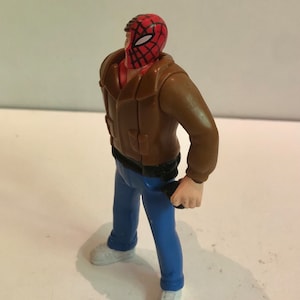 May include: A plastic action figure of a man wearing a brown jacket, blue jeans, and a red and black spider-man mask.