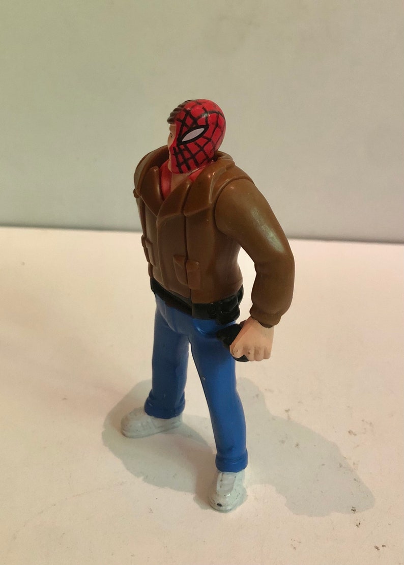 May include: A plastic action figure of a man wearing a brown jacket, blue jeans, and a red and black spider-man mask.