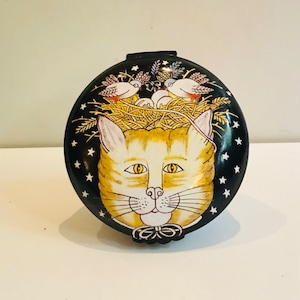 May include: A round, black trinket box with a white cat face and a nest of birds on top. The cat has yellow eyes and a black bow tie. The background is black with white stars.
