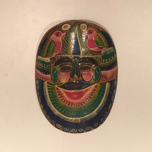 May include: A colorful wooden mask with a smiling face, painted with blue, green, pink, and yellow. Two birds are painted on the top of the mask.