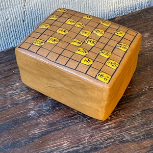 以下が含まれることがあります： 茶色の仕上げをした小さな長方形の木箱。箱の上部はグリッドパターンと黒い線で飾られています。グリッドの正方形には黄色の中国語の文字が書かれています。
