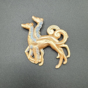 Pode incluir: Um broche dourado com dois galgos estilizados com detalhes azuis. Os galgos estão olhando para a frente com as caudas enroladas.