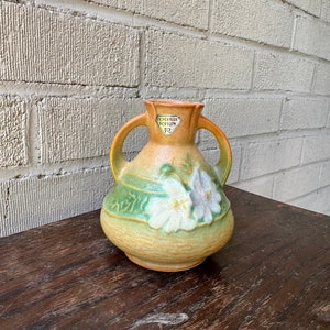 May include: A green and brown glazed ceramic vase with white floral designs and two handles. The vase has a label that reads "Roseville Pottery 12".
