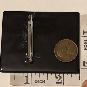 May include: Black square brooch with a silver pin on the back. The brooch is about 1 inch wide and 1 inch tall.