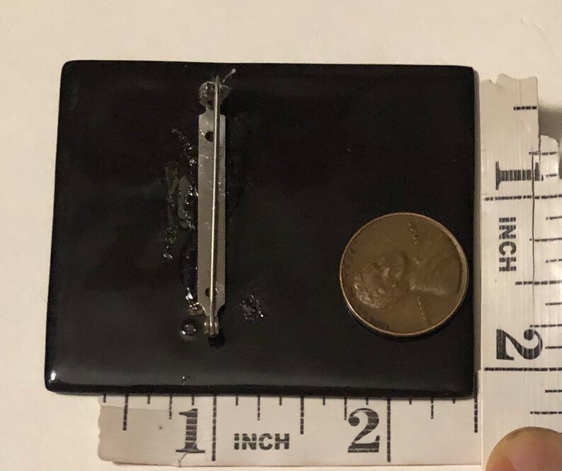 May include: Black square brooch with a silver pin on the back. The brooch is about 1 inch wide and 1 inch tall.