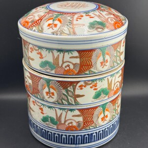 May include: A cylindrical ceramic stacking container with a lid, featuring a white base decorated with intricate patterns in orange, green, and blue. The design includes stylized floral motifs, pine trees, and a geometric blue border at the base.