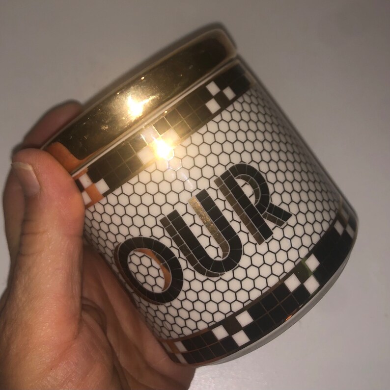 May include: White ceramic jar with a gold lid and black and gold geometric pattern. The word "OUR" is written in gold letters on the front of the jar.