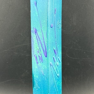 Puede incluir: Un jarrón de cerámica alto y rectangular con una abertura superior inclinada. La superficie presenta un degradado de azul turquesa brillante que transiciona a una base gris metálico oscuro, con líneas púrpuras texturizadas y símbolos en relieve.