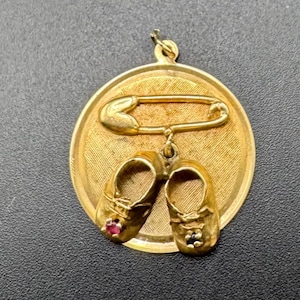 May include: A gold-toned pendant featuring a safety pin design with a pair of small baby shoes hanging below. One shoe has a small red gem. The pendant is round with a textured surface and a loop at the top for a chain.